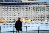 A man stands in the foreground of a shot of a large cruise ship with people on the balconies