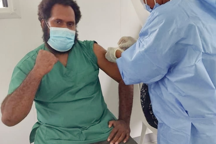 A man wearing a green shirt and mask holds his fist in the air as a person in scrubs leans over his arm.