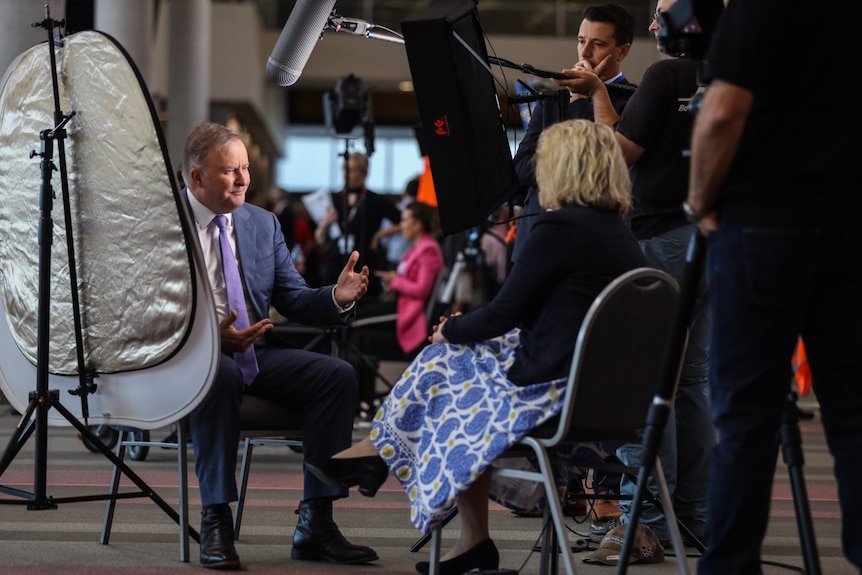 Anthony Albanese sits in a chair opposite a reporter, with a boom microphone overhead and a reflector bouncing light onto his fa