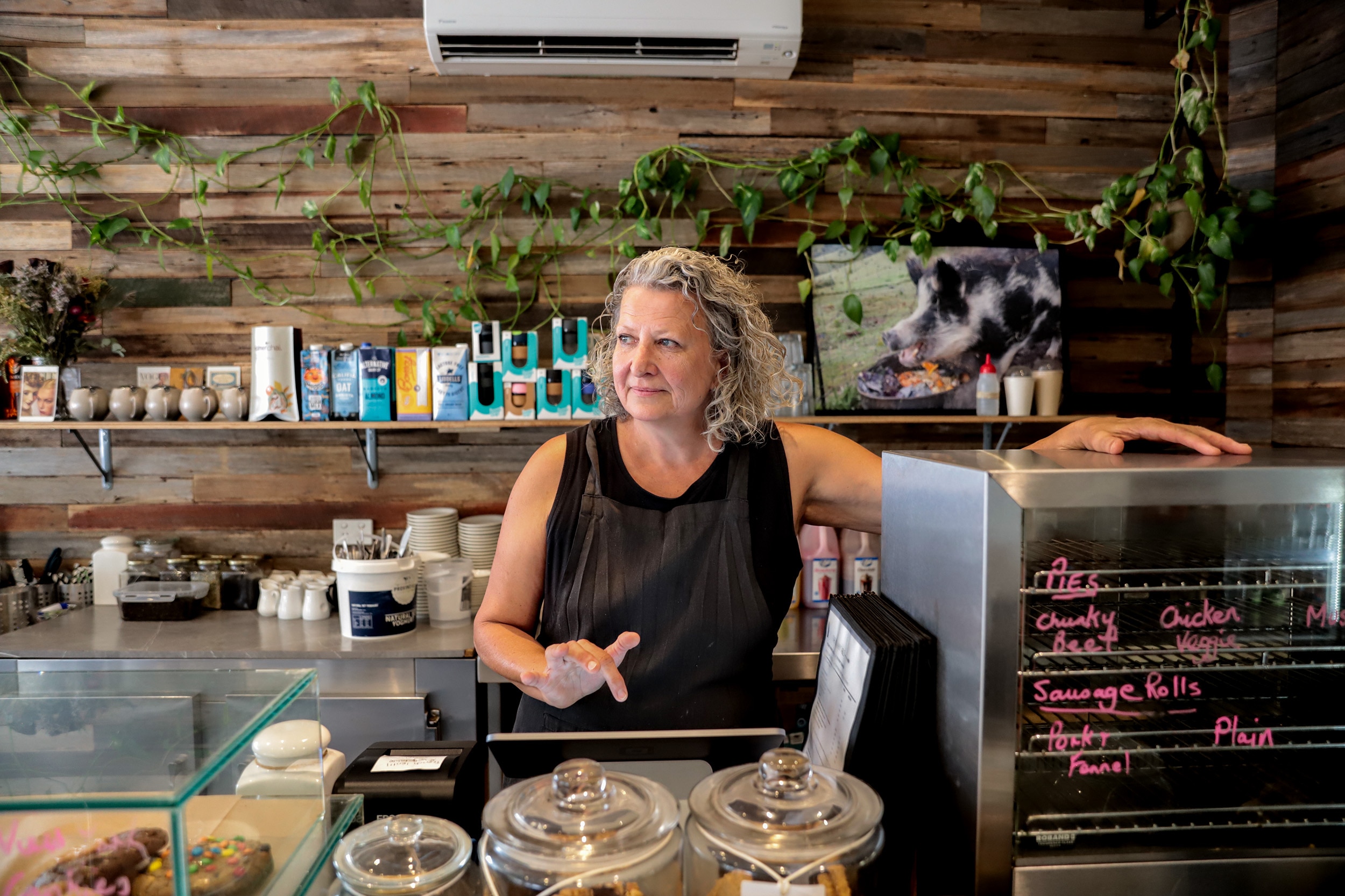 Woman with grey hair and black top stands behind cafe counter with hand over register looking out towards window 
