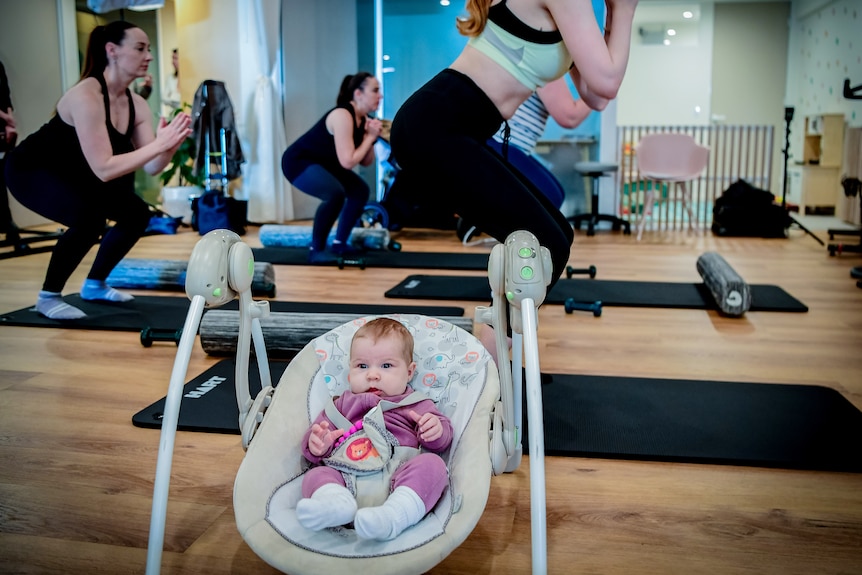 A baby lies in a cradle while women wearing active gear do squats duirng an exercise class