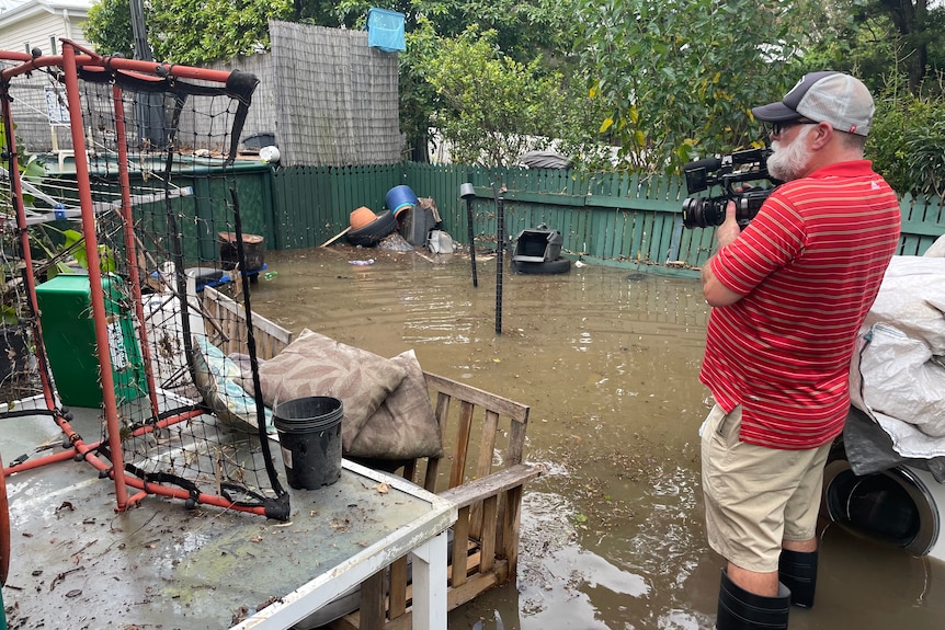 Man wearing gumboots, standing in flooded water and filming with a camera.