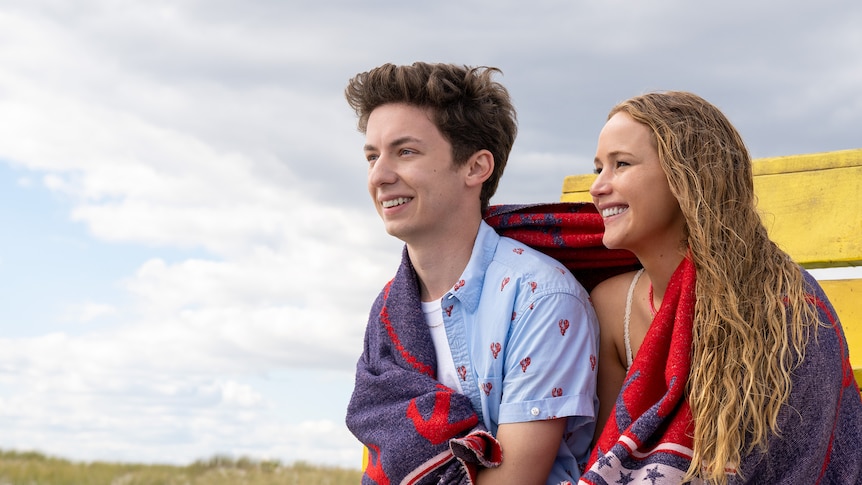 A white teen boy with mousey brown hair and blue shirt sits wrapped in a beach towel with a white woman with blonde wavy hair