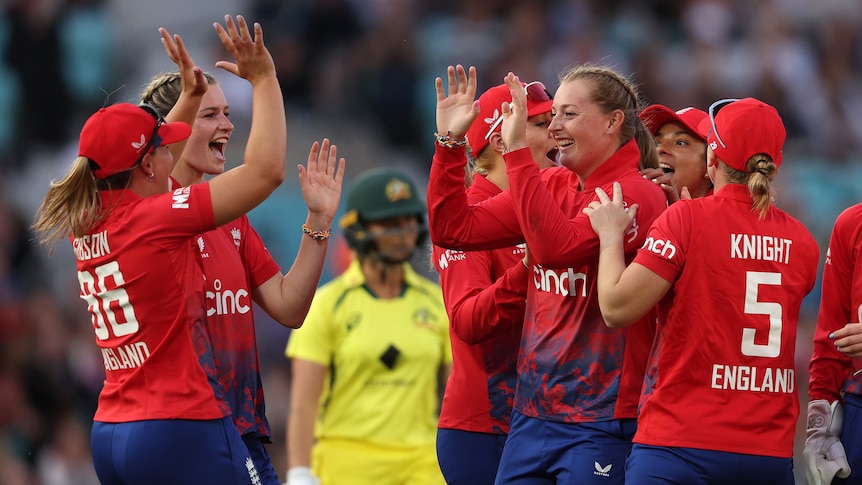 A group of excited English women cricketers high-five and celebrate as a dejected Australian batter stands in the background.