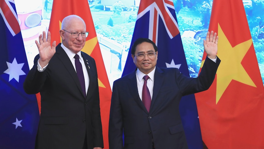 A man waving next to a Vietnamese man in maroon tie. Both wearing suits and standing in front of Australian and Vietnamese flags