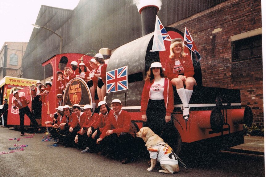 A group of people are gathered around a red steam train with a Union Jack on it. Elaine is at the front with her guide dog.