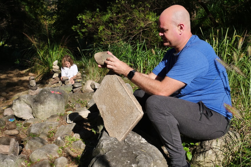 A man in the foreground place a small rock on a large paver while a boy sits in the background among rocks.