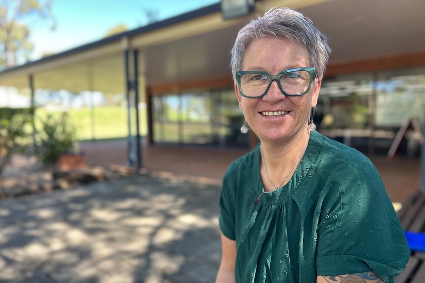 A smiling woman with short grey hair, green shirt and green glases sitting outside a small business.