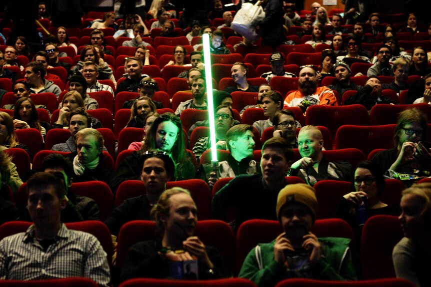 A Star Wars fan sits in a cinema holding his lightsaber surrounded by other Star Wars fans.
