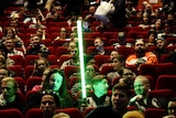 A Star Wars fan sits in a cinema holding his lightsaber surrounded by other Star Wars fans.