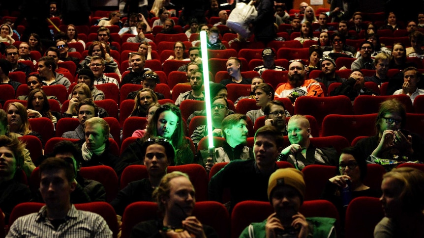 A Star Wars fan sits in a cinema holding his lightsaber surrounded by other Star Wars fans.