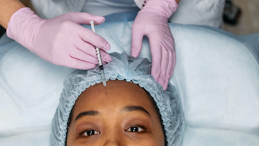 A woman, wearing a shower cap, has botox injected into her forehead.