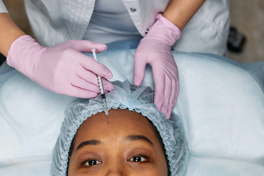 A woman, wearing a shower cap, has botox injected into her forehead.