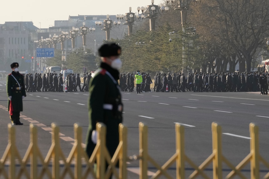 Security guards line a closed street as crowd of people gather in the distance. 