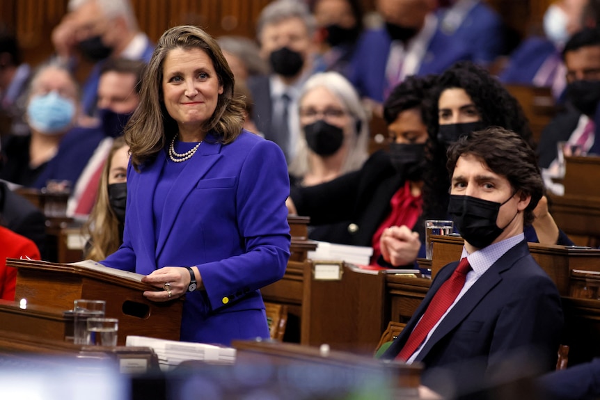 Freeland se encuentra en un atril en el Parlamento.  Trudeau se sienta cerca, con una máscara.  Ambos miran más allá de la cámara.