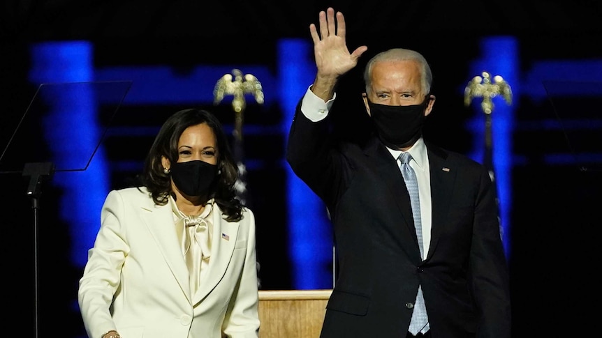 Joe Biden waves, next to Kamala Harris on stage in front of an American flag.