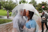 A couple wearing plastic ponchos kiss under a plastic umbrella.