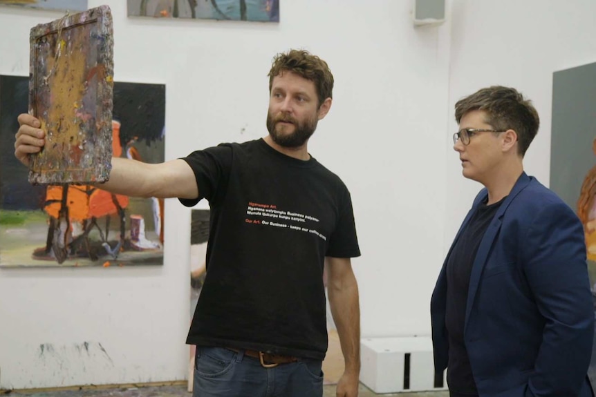 Ben Quilty and Hannah stand talking in his studio while he holds a small mirror up to demonstrate how he paints self-portraits.