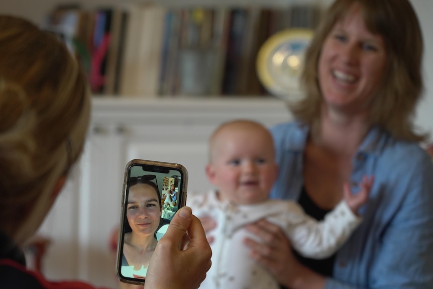 A woman holds a baby while being filmed on an iPhone for a Facetime call
