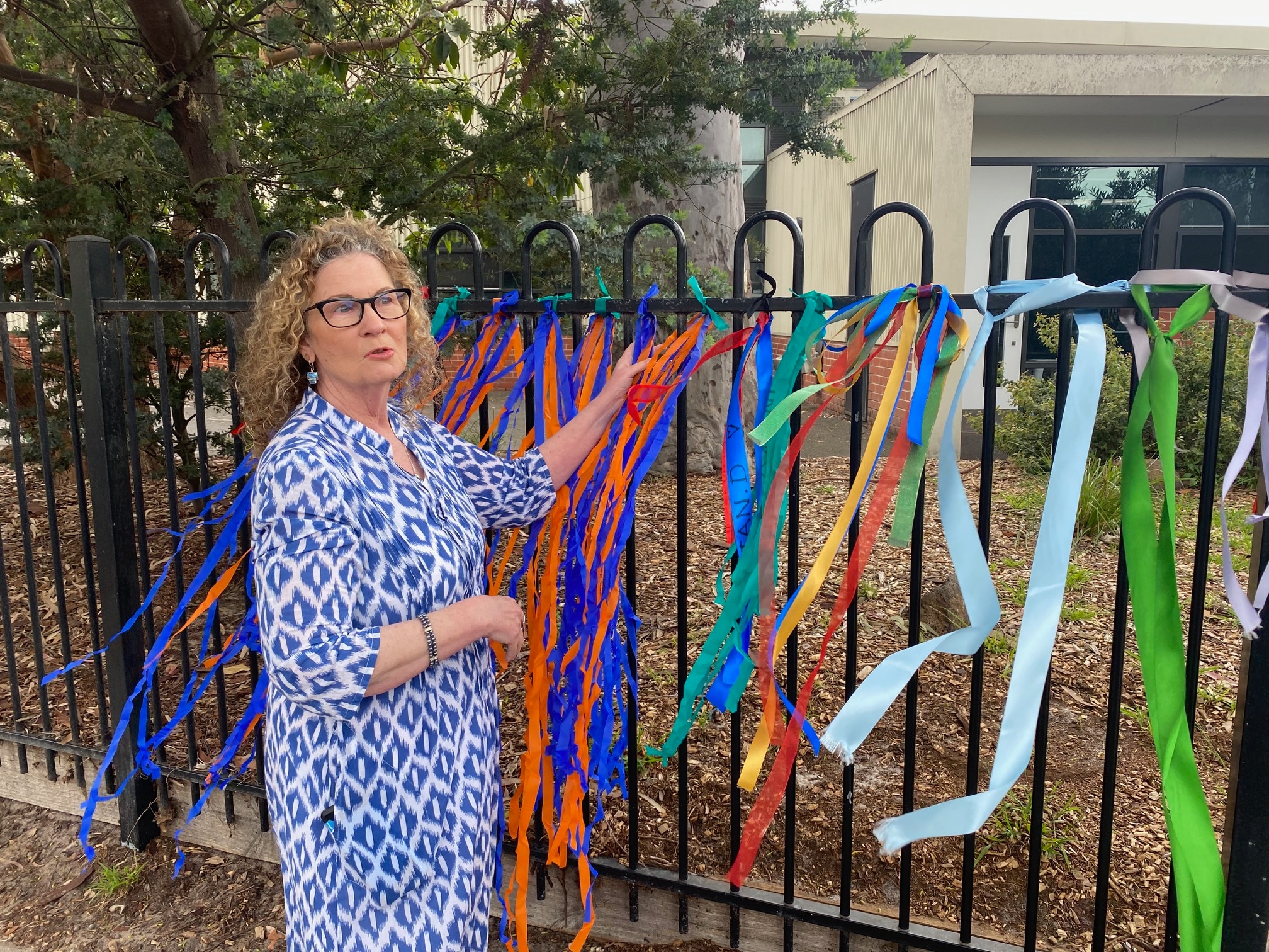 A woman in a blue and white dress stands next to a fence to which colourful ribbons are tied.