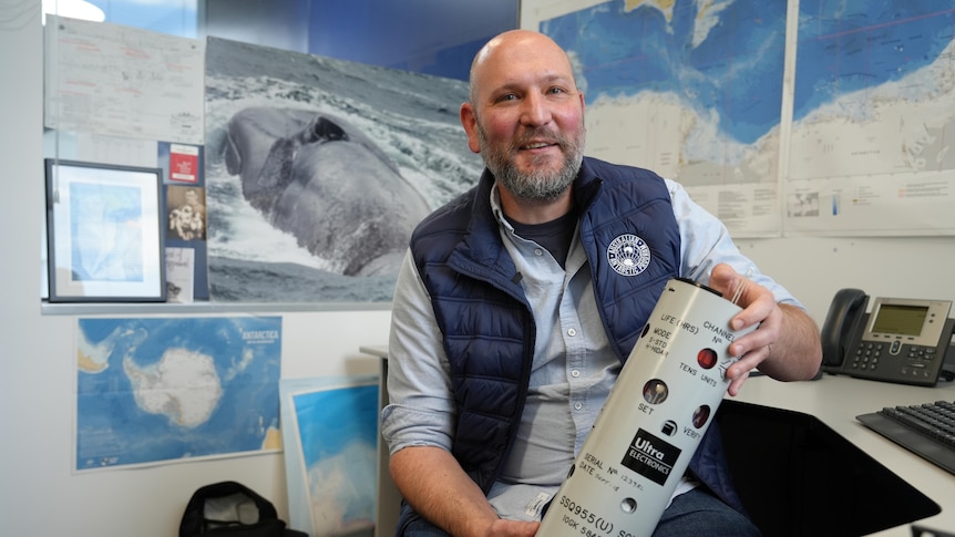 Dr Brian Miller in his office holding a medium-sized cylinder with pictures of whales and maps behind him