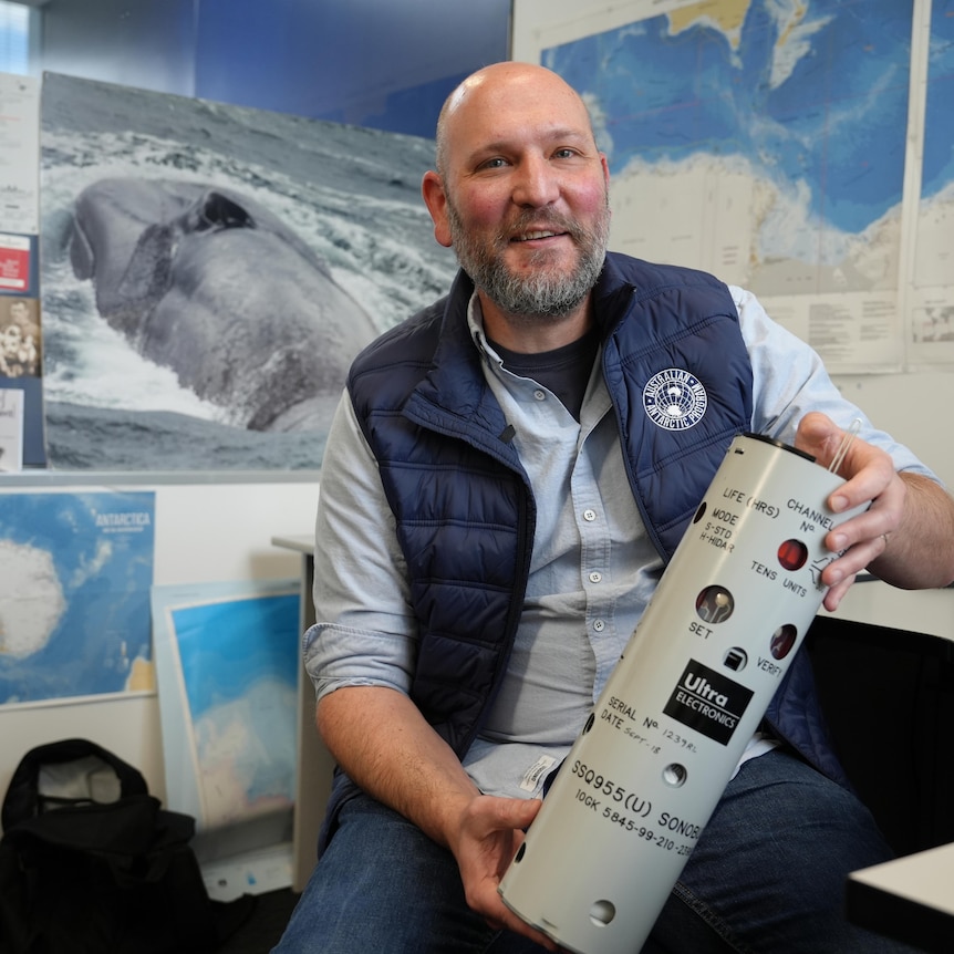Dr Brian Miller in his office holding a medium-sized cylinder with pictures of whales and maps behind him