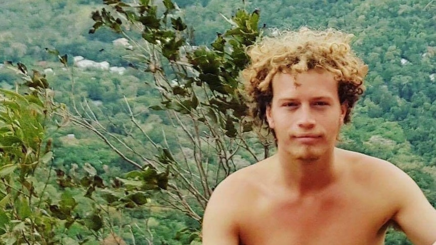 A young, shirtless man with curly blonde hair stands in front of a bushy valley.