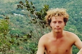 A young, shirtless man with curly blonde hair stands in front of a bushy valley.