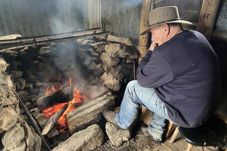 A man sits next to a fire in an outdoor shed wearing a wide brim hat, a blue jumper and jeans.