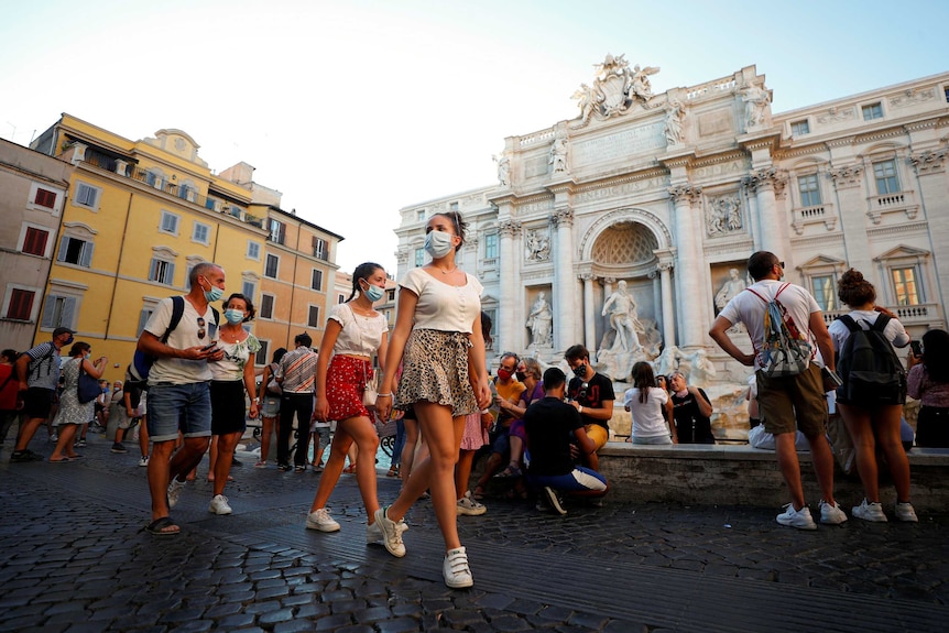 Two young women, followed by two adults, in face masks, walk past ornate buildings on summer's day.