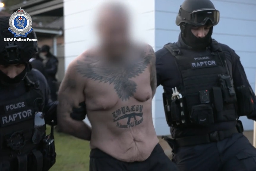 A shirtless man with his hands behind his back flanked by police in helmets and vests