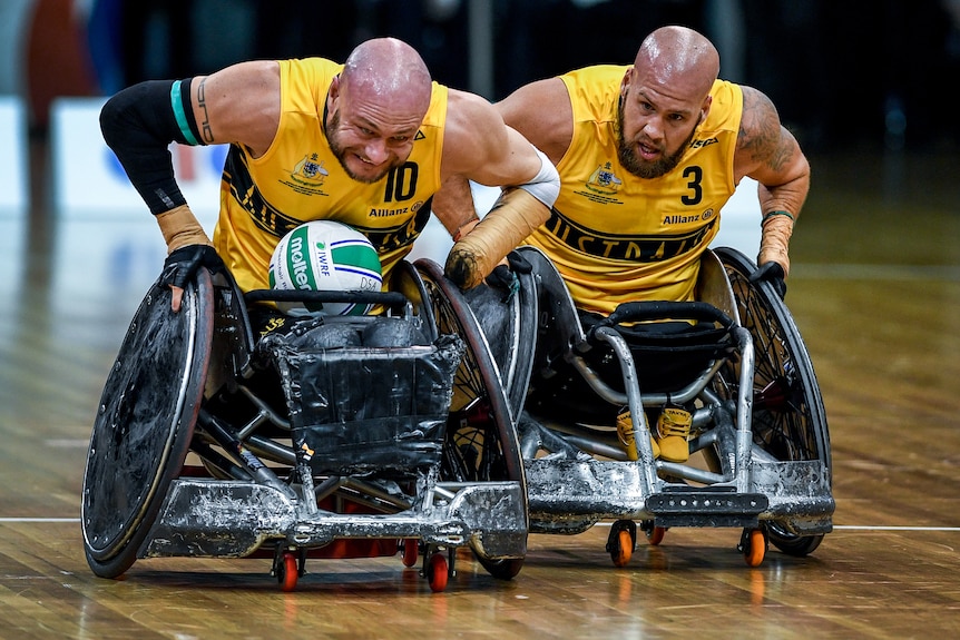 Two Australian wheelchair rugby players push their chairs down the court, with one holding the ball in his lap.