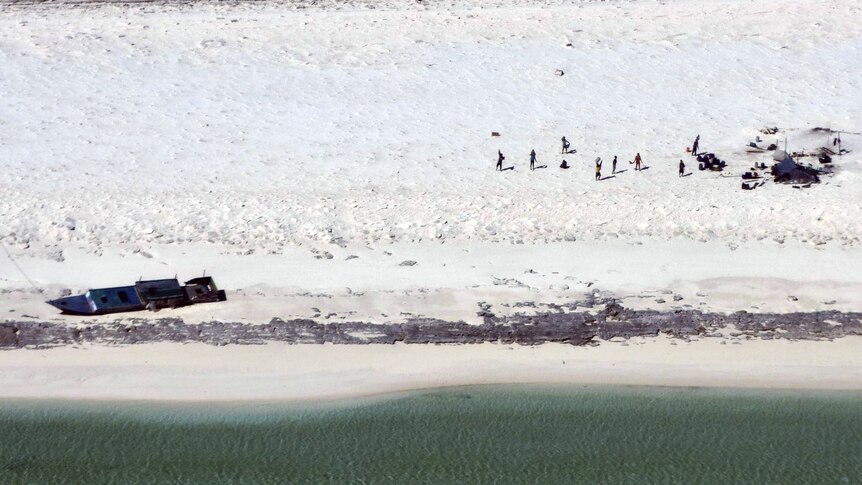 A sandy beach with a washed up boat and eight people visible near a makeshift camp. 