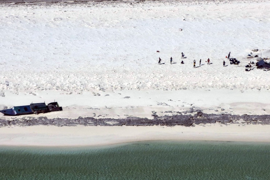 A sandy beach with a washed up boat and eight people visible near a makeshift camp. 