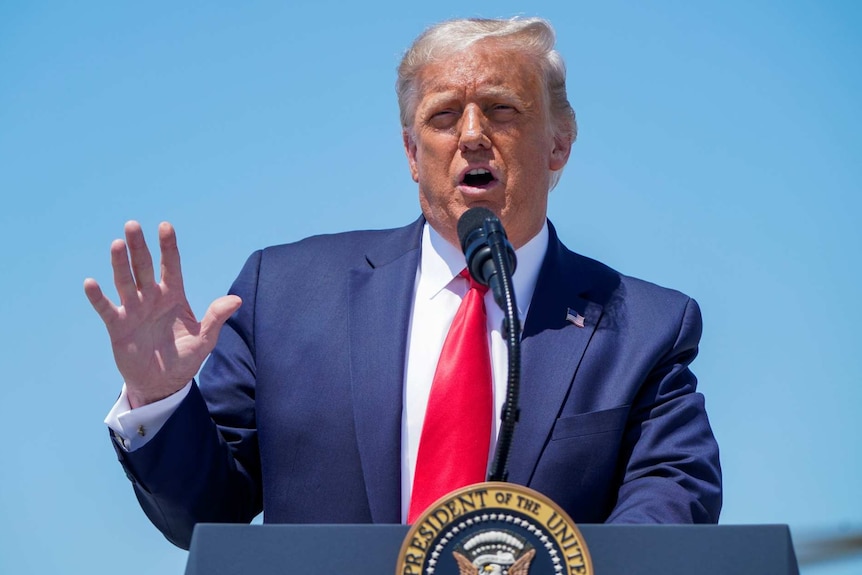 donald trump wearing a suit and red tie at a microphone holding up his right arm as he speaks with daylight sky behind him