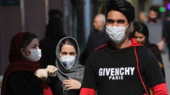 A man wearing a face mask walks past two women also wearing masks on a cool and sunny day.