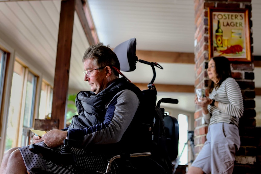 Man with glasses in wheelchair holding a letter with woman behind him next to brick column inside house holding a mug