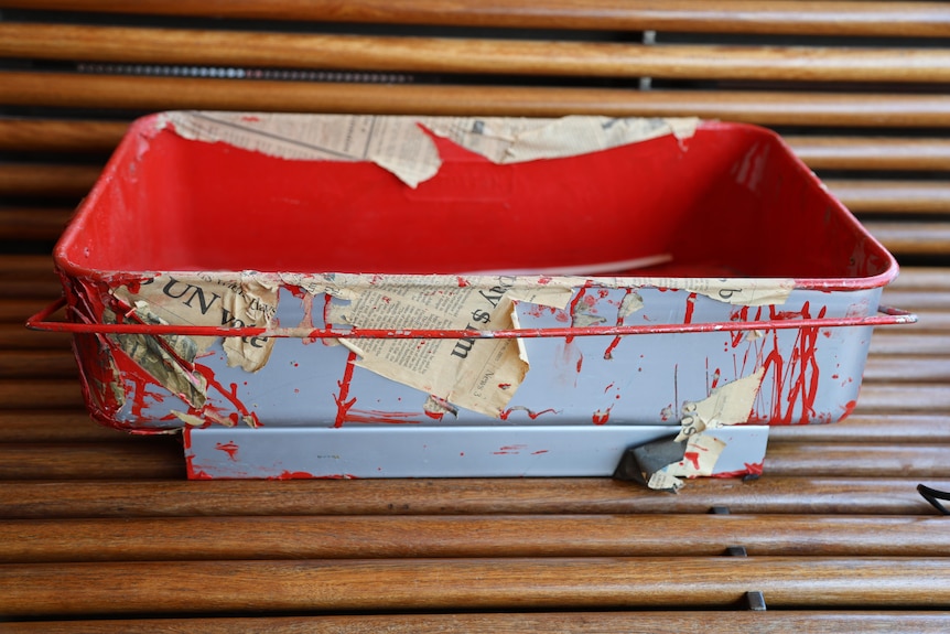 A large rectangular paint tray sits on a park bench. It is stained with thick red paint and remnants of yellowing newspaper