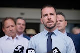 A man in a collared shirt and tie speaking outside of a courthouse, with several other men in the background.