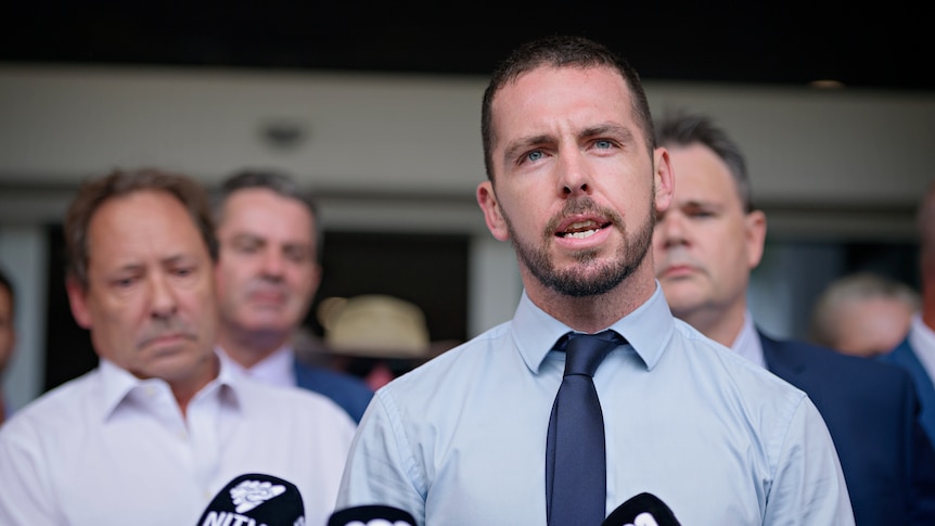 A man in a collared shirt and tie speaking outside of a courthouse, with several other men in the background.