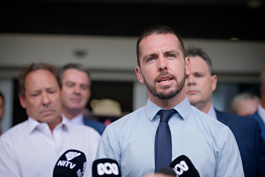 A man in a collared shirt and tie speaking outside of a courthouse, with several other men in the background.