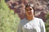 A young women standing and looking serious, standing outside on a sunny day in front of red rocks and greenery.