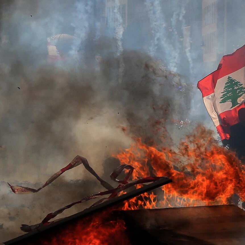 smoke filled street with fire in front. Figure carries Lebanese flag