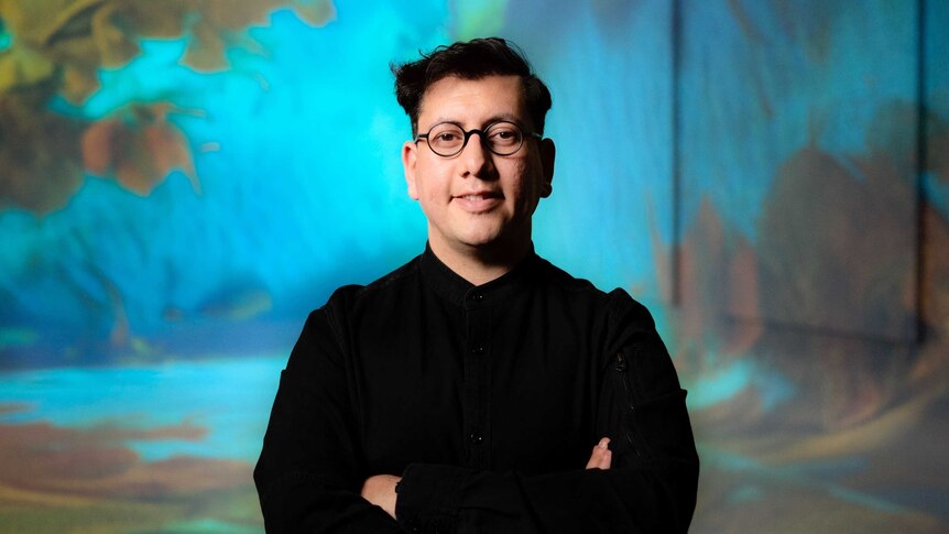 A dark haired man with a friendly face and glasses stands with his arms crossed. he wears a black shirt.