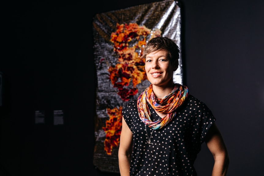 Anna Madeleine Raupach smiles in front of her Ramsay Art Prize entry, a red and orange embroidered work