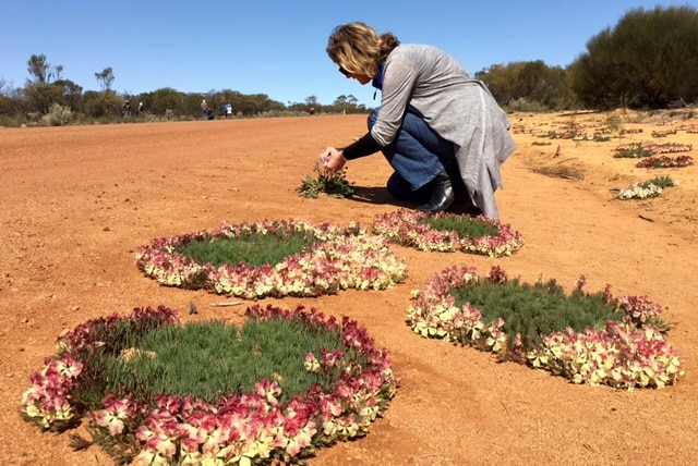 A woman crouches by the side of a dirt road to look at a pink wreath flower.