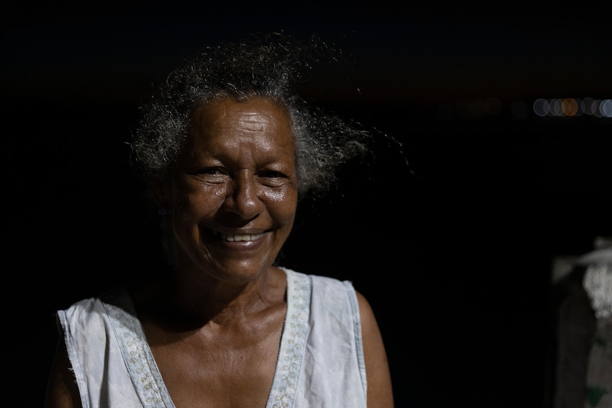 Enid Tom, with grey hair and a wide smile, is against a dark background, as if it's nighttime.