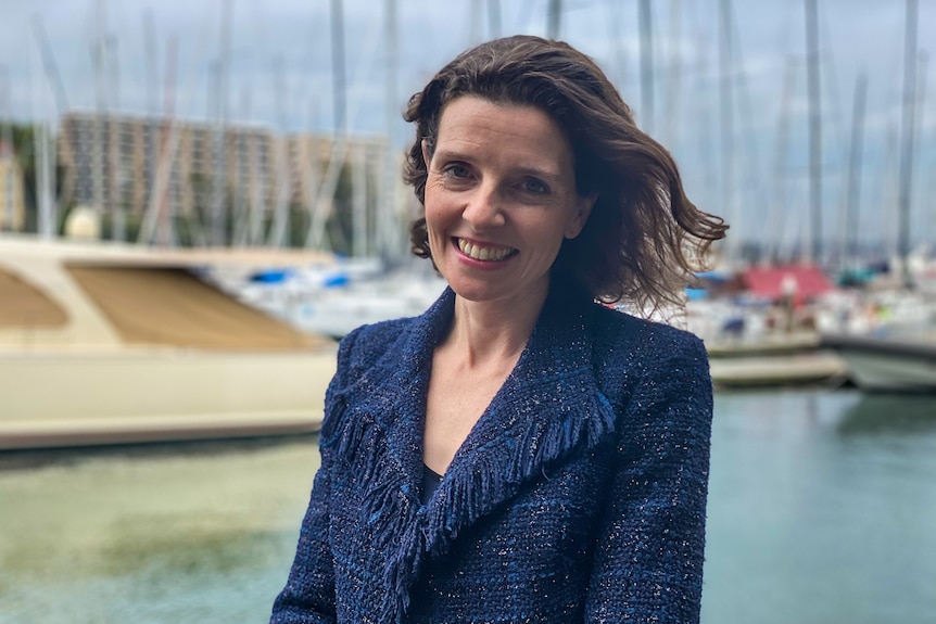 a woman standing outdoors near a marina, smiling. She is wearing a dark blue suit and the wind is in her hair