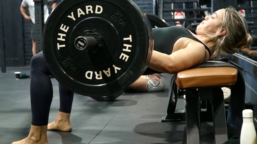 A woman lies on a weight bench preparing to lift a set of weights with "The Yard" written on them in white letters.
