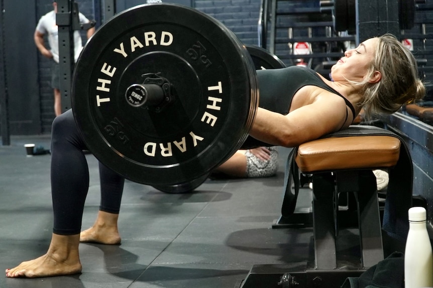 A woman lies on a weight bench preparing to lift a set of weights with "The Yard" written on them in white letters.